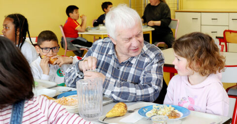 Une photo montrant un senior souriant mange à la table des écoliers avec des enfants à ses côtés dans une cantine à Cergy